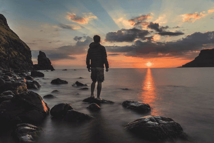 person a sunset standing on a rock by water